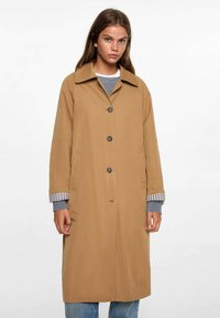 Cappotto lungo marrone con colletto, chiusura a bottoni frontale e tasche laterali. Le maniche presentano un motivo a quadretti e il tessuto appare liscio e leggero.