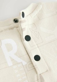 Tkanina w kolorze kremowym z subtelnymi wzorami, z czarnymi zatrzaskami i teksturowaną powierzchnią. Projekt zawiera wyraźne akcenty tekstowe.