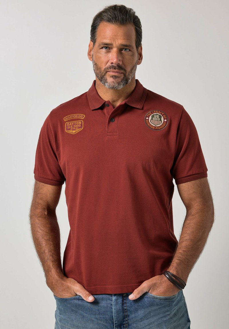 Hombre con barba que lleva una camiseta polo granate con parches de aviación, manos en los bolsillos y vaqueros azules sobre un fondo liso.