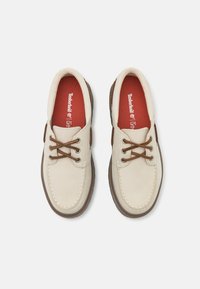 Sapatos de couro bege claro com acabamentos castanhos, bico redondo e atacadores castanhos. Palmilhas vermelhas com o logótipo "Timberland". Sola de borracha com textura para melhor aderência.