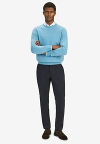 Ljusblå crew-neck-tröja av stickad tyg, i kombination med mörka byxor. Modellen står med armarna i kors, iförd bruna skor.