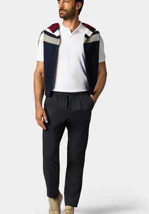 Marine mouwloze vest met rode, grijze en witte strepen over een witte poloshirt, gecombineerd met donkere broeken en beige sneakers.