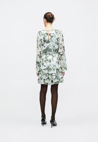 Vrouw met haar in een knot, gekleed in een groene en witte bloemenjurk, zwarte panty's en zwarte hoge hakken, staand met de rug naar de camera tegen een effen achtergrond.