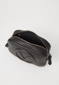 Svart läder crossbody-väska med en texturerad yta, guldfärgad dragkedja och präglad cirkulär logotyp på framsidan. Liten dragkedjeficka inuti.