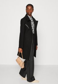 Abrigo de lana negro con un cierre asimétrico, combinado con una blusa de estampado de cebra y pantalones negros. Sostiene un pequeño bolso beige.
