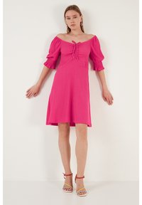 Pink, off-the-shoulder dress with puffed sleeves and a front tie detail. Flared skirt with a textured fabric. Model wearing multicolored heels.