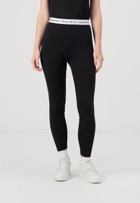 Črne legice iz raztegljive tkanine z belim pasom, na katerem je napis "Tommy Jeans" in rdeči poudarki. Oblikovan kroj, do gležnjev.