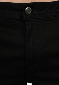 Pantaloni in denim nero con chiusura a bottone metallico e cuciture a contrasto. La vista ravvicinata mostra la texture e i dettagli delle cuciture attorno alla cintura.