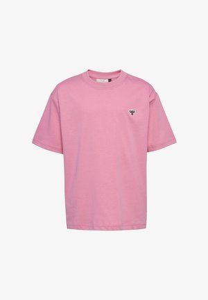 Młoda dziewczyna stoi, ubrana w różowy, luźny t-shirt i dresy, białe trampki, uśmiecha się na jednolitym jasnym tle.