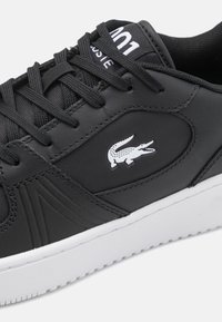 Schwarze Sneakers aus synthetischem Leder mit weißer Gummisohle. Verfügt über strukturierte Seitenpaneele und ein kleines weißes Krokodil-Logo auf der Außenseite.