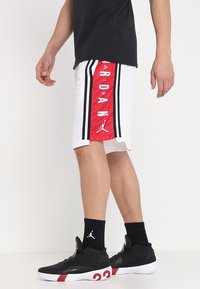 Personne portant un short de basketball Jordan blanc et rouge avec des rayures noires, des chaussettes Jordan noires et des chaussures de basketball noires avec le numéro rouge 23.