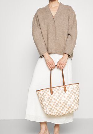 Person i beige tröja och vit kjol som håller en beige och vit rutig shoppingväska med bruna handtag.