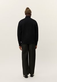 Svart sweatshirt med hög krage, kombinerad medvida mörkgrå byxor och en brun mössa. Slät tyg med avslappnad passform.