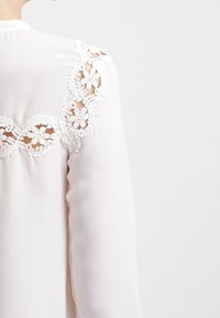 Blouse blanche à manches longues, en tissu lisse, avec une bordure en dentelle décorative le long des épaules et des motifs floraux.