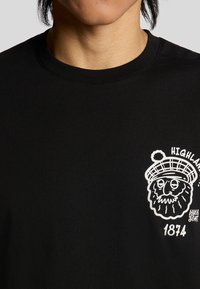 Sort bomulds T-shirt med et hvidt grafik af en skægget figur i en hat, med teksten "HIGHLAND" og "1874". Rund hals, korte ærmer.