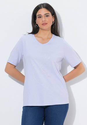 Joven con cabello largo y oscuro que lleva una camiseta de cuello en V color lavanda clara con un diseño sutil de un gato, combinada con jeans azules, parada con las manos detrás de la espalda.