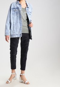 Jeansjacke in hellblau, olivgrünes Tanktop mit grafischen Details, schwarze, schmal zulaufende Hose mit unterschiedlicher Textur und silberne Blockabsatzsandalen.