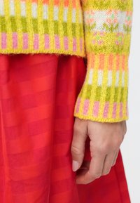 Pletený sveter so žltými, ružovými a zelenými pruhmi vrstvený cez červenú sukňu s kockovaným vzorom; ruka spočíva na okraji sukne.