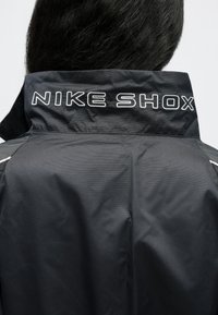 Μαύρο μπουφάν Nike Shox με ψηλό γιακά, ραβδωτή υφή και το κέντημα "NIKE SHOX" σε λευκό χρώμα στην πλάτη.