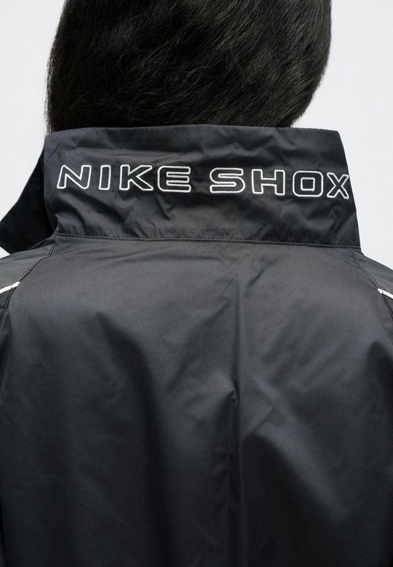 Schwarze Nike Shox Jacke mit hohem Kragen, geripptem Texturdesign und "NIKE SHOX" in Weiß auf dem Rücken bestickt.