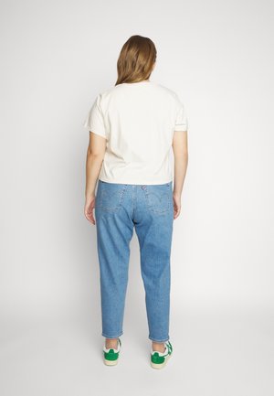 Personne debout de dos, portant une chemise blanche à manches courtes, un jean bleu et des baskets blanches avec des accents verts sur fond blanc.