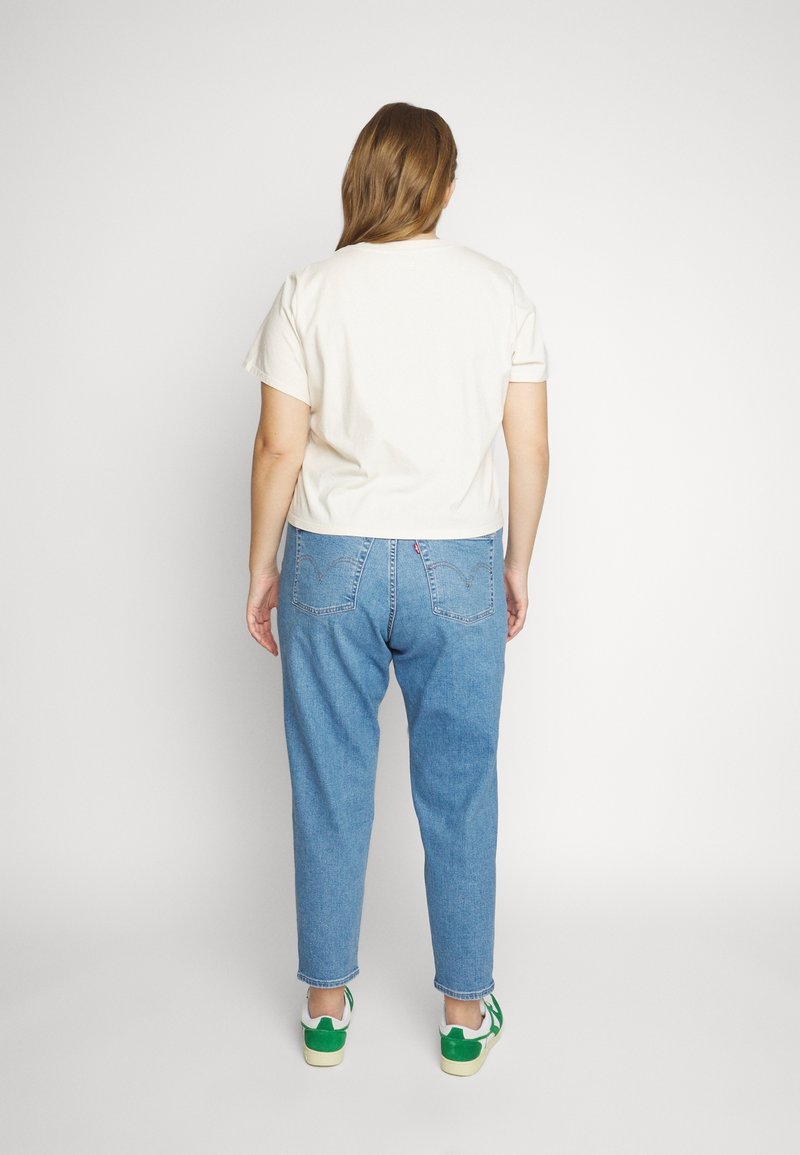 Personne debout de dos, portant une chemise blanche à manches courtes, un jean bleu et des baskets blanches avec des accents verts sur fond blanc.