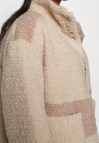 Flauschige beiger Jacke mit hohem Kragen, ausgestattet mit pinkfarbenen strukturierten Patches und einer Reißverschlusstasche. Weicher, taktiler Stoff mit boxy Silhouette.