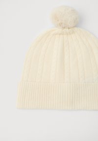 Krémová čepice s pleteným vzorem a ohrnutým ribbed okrajem, na vrcholu s nadýchaným pom-pomem. Vyrobena z měkkého, texturovaného materiálu.