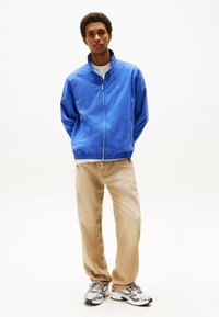 Giacca blu con zip, colletto a imbuto, polsini e vita elasticizzati, abbinata a pantaloni beige e scarpe da ginnastica argentate.