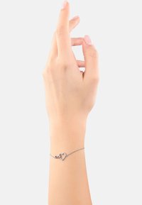 Mano con un tono di pelle chiaro che indossa un delicato braccialetto d'argento con un charm a forma di cuore, su uno sfondo bianco semplice.