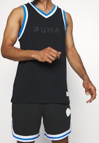 Man som bär svart Puma ärmlös sporttröja och matchande shorts med blå och vit trim, stående mot en plain bakgrund.