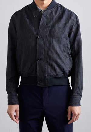Homme portant une veste bomber bleu marine texturée à boutons et un pantalon foncé assorti sur un fond clair uni.