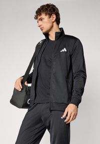 Black athletic track jacket with zip closure and high collar, featuring white logo accents. Smooth fabric design, paired with matching pants and bag.