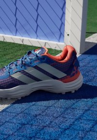 Sininen ja oranssi urheilukenkä, jossa valkoiset raidoitukset, sijoitettuna sinivalkoiselle nurmikentälle valkoisen urheilutolpan lähelle.