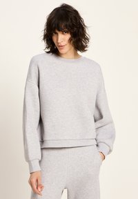 Frau mit kurzen, lockigen, dunklen Haaren, die einen hellgrauen Sweatshirt und passende Jogginghosen trägt, steht mit einer Hand in der Tasche vor einem schlichten Hintergrund.