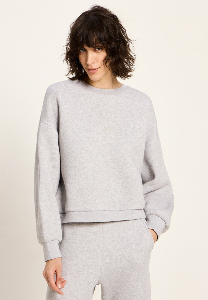 Frau mit kurzen, lockigen, dunklen Haaren, die einen hellgrauen Sweatshirt und passende Jogginghosen trägt, steht mit einer Hand in der Tasche vor einem schlichten Hintergrund.