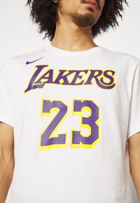 Bílá bavlněná tričko s nápisem "LAKERS" v purpurových a žlutých písmech, s číslem "23" v purpurové barvě s žlutým orámováním. Logo Nike na rameni.