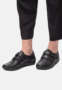 Zwarte leren schoenen met dubbele verstelbare banden, ronde neus, gestructureerd oppervlak en flexibele zool, gedragen met cropped zwarte broeken.