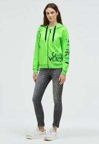 Frau steht und trägt eine hellgrüne Kapuzenjacke mit Reißverschluss und schwarzem Text, dunkelgraue Skinny-Jeans und weiße Turnschuhe vor einem schlichten hellen Hintergrund.