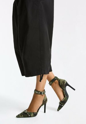 Jambes en pantalon court noir portant des talons hauts camouflage verts avec brides à la cheville et clous en métal sur fond blanc.
