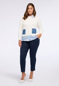 Femeie stând în picioare, purtând un pulover alb cu buzunare bleumarin peste o cămașă bleumarin deschis, blugi bleumarin închis și pantofi cu toc alb pe un fundal simplu.