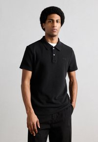 Jeune homme avec coiffure afro portant un polo à manches courtes noir et un pantalon noir, debout devant un fond clair uni.