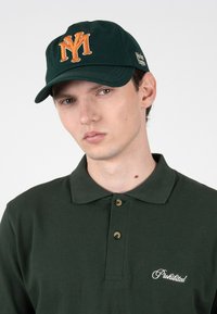 Jeune homme portant un polo vert foncé avec l’inscription "Prohibited" brodée et une casquette noire avec un logo "NY" orange et un texte latéral blanc.