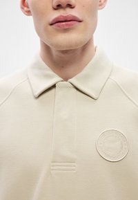 Texturált anyagból készült bézs póló, rövid, hegyes nyakkal, és bal oldalon, a mellkason emelt teniszező klub logóval.