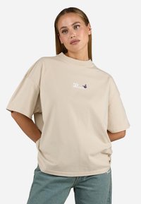 T-shirt in cotone oversized beige con maniche corte, caratterizzata da testo ricamato e un design di farfalla viola sul petto.