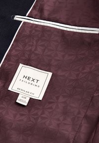 Pruunika toonides mustriga kangas, millel on geomeetriline disain ja tekstuuriga pind; sisaldab valget silti kirjaga "NEXT Tailoring" ja suuruste andmed.