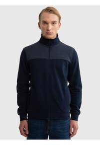 Jonge man met blond haar, gekleed in een marineblauwe jas met rits en een donkergrijs paneel over de schouders, staand tegen een effen achtergrond.