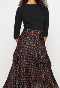 Czarny dopasowany top z rękawami trzyczwartowymi, w połączeniu z długą, wzorzystą spódnicą z czerwono-białymi geometrycznymi wzorami i szczegółem w postaci zawiązanego pasa.