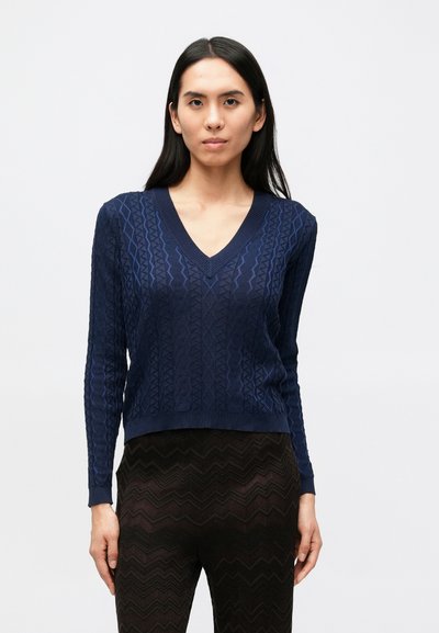 Pull tricoté bleu marine avec un col en V et des manches longues. Présente un motif texturé avec des formes en zigzag. Porté avec des leggings foncés à motif.