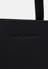Svart syntetisk tote-väska med präglad "CALVIN KLEIN JEANS"-logotyp, slät yta och robusta remmar. Minimalistisk design med rena linjer.